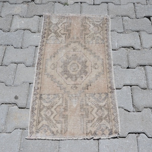 May include: A rectangular woven rug featuring a central geometric medallion pattern in shades of beige, tan, and muted brown. The textile displays symmetrical motifs and borders with a short fringe along the top and bottom edges of the piece.