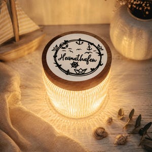 Op de afbeelding: Een cilindrische glazen pot met verticale ribbels en een warme interne gloed. De pot heeft een rond houten deksel met een witte inzet aan de bovenkant. De inzet toont de zwarte tekst "Heimathafen" omringd door nautische iconen, waaronder een anker, een scheepsroer, een papieren bootje en vogels.