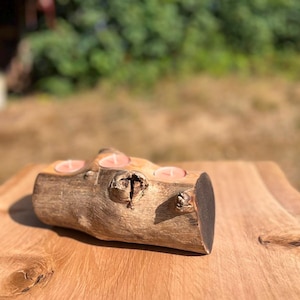 May include: A natural wooden log candle holder featuring a rough, textured bark exterior with visible knots and three circular indentations along the top, each containing a small, round, light pink tea light candle with a white wick.