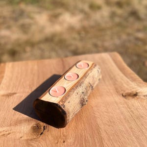 May include: A cylindrical wooden log candle holder featuring a flat top surface with three circular cutouts. Each cutout contains a small, round, light pink tea light candle with a centered white wick. The log retains its natural bark texture.