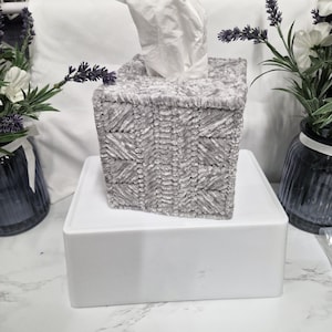 May include: A square tissue box cover featuring a textured, light grey, chenille-style yarn with a repeating geometric pattern of diagonal lines. A single white facial tissue is pulled through the top opening of the box cover.