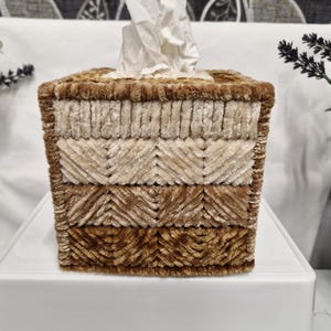 May include: A square tissue box cover featuring a textured, multi-layered design in shades of brown and cream. The exterior is crafted from soft, plush chenille-like yarn arranged in horizontal bands with alternating chevron and straight patterns.