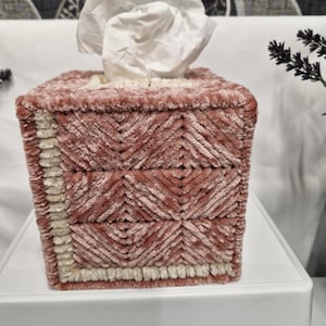 May include: A square tissue box cover crafted from textured, dusty rose velvet yarn. The design features a geometric chevron pattern across the sides, accented by a vertical and horizontal border of cream-coloured yarn along the front edge.