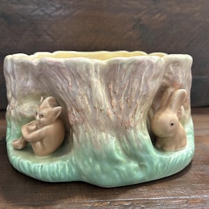 May include: A ceramic planter shaped like a tree stump with a scalloped rim. The exterior features a textured bark pattern in shades of pink and brown, with a green base. A small sculpted figure of a seated imp and a rabbit are attached.