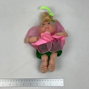 May include: A doll with blonde hair tied with a light green ribbon, green eyes, and freckles on its cheeks. It wears a pink glittery wing-like garment, a pink fabric flower bodice, and dark green shorts. A ruler shows it is 23 cm tall.