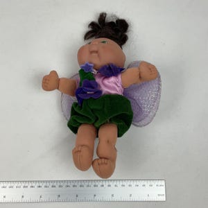 May include: A doll with dark brown hair, a light pink top, and a dark green skirt featuring purple flower accents. The doll has light purple glittery wings and is positioned above a ruler showing measurements in inches and millimetres.