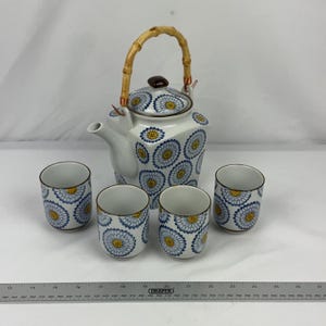 May include: A white ceramic teapot with a bamboo handle and three matching cups, all decorated with a blue and yellow floral pattern. A metal ruler is placed in front, showing the teapot is approximately 18 cm tall and 15 cm wide.