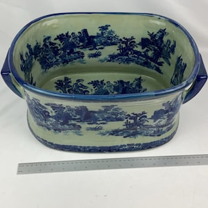 May include: An oval ceramic basin with a cream-coloured base and blue transferware patterns depicting pastoral scenes. The basin features two side handles and a blue rim. A metal ruler is placed in front, showing a length of 40 cm.