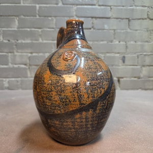 May include: A ceramic jug with a rounded body and a narrow neck, featuring a glossy glaze in shades of brown and black. The surface displays a mottled texture with dark, swirling patterns and a small, circular embossed detail near the top.