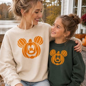 May include: A beige crewneck sweatshirt and a dark green crewneck sweatshirt, both featuring a large orange graphic of a pumpkin shaped like a mouse head with a smiling face. The sweatshirts have long sleeves and ribbed cuffs, made of a soft fabric.
