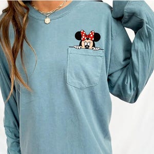 May include: A long-sleeve, dusty blue crewneck shirt featuring a chest pocket. A graphic of a cartoon mouse with a red and white polka-dot bow is positioned so that the character appears to be peeking out from the top edge of the pocket.