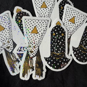 May include: A collection of die-cut stickers featuring two distinct designs: a white figure with black polka dots, a long nose, and a gold triangle, and a black figure with white polka dots, a long nose, and gold accents, all with holographic details.