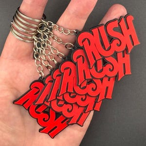 May include: A collection of multiple keychains featuring the word RUSH in bold, bright red lettering with a black outline. Each keychain is attached to a silver-colored metal split ring by a short metal chain, creating a layered, stacked appearance.