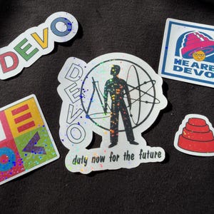 May include: A collection of five DEVO stickers with holographic finishes. One sticker features the text DEVO, another shows a figure with a beaker and atom, one displays the text WE ARE DEVO, one is a square grid, and one is a red energy dome.
