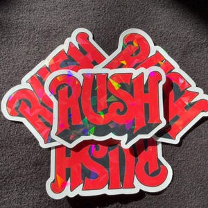May include: A collection of die-cut stickers featuring the word RUSH in bold, red, stylized lettering. The letters have a holographic, multi-colored geometric pattern that reflects light, and each sticker has a thick white border outline.