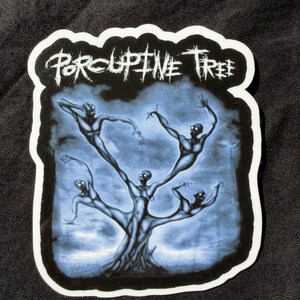 May include: A die-cut sticker featuring the text Porcupine Tree in a jagged white font at the top. Below the text, a dark blue and black illustration depicts a tree-like figure composed of four humanoid shapes with glowing white eyes.