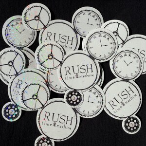 May include: A collection of circular white stickers featuring black clock face designs and the text RUSH time machine. Some stickers have a holographic finish, while others display gear motifs with the number 3 in the center of the gears.