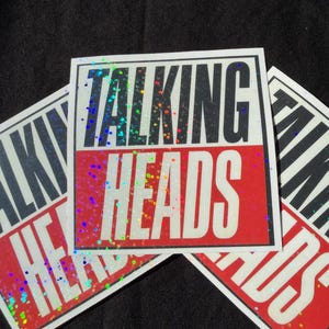 May include: A square sticker featuring the text TALKING HEADS in a bold, two-tone design. The word TALKING is printed in black on a white background, while the word HEADS is in white on a red background. The entire surface has a holographic glitter finish.