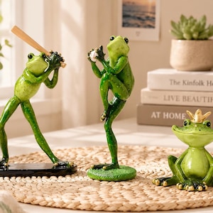 Peut inclure: Trois figurines de grenouilles vertes : une debout sur une base noire tenant une batte de baseball en bois, une debout sur une base verte attrapant une balle de baseball blanche, et une assise portant une petite couronne dorée.