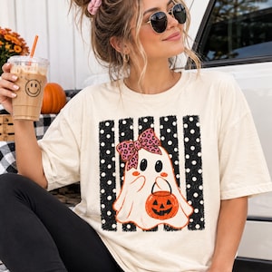 May include: A cream-colored short-sleeve t-shirt featuring a graphic print of a white ghost wearing a pink leopard print bow and holding an orange jack-o'-lantern bucket. The ghost is centered over a background of black vertical stripes with white polka dots.