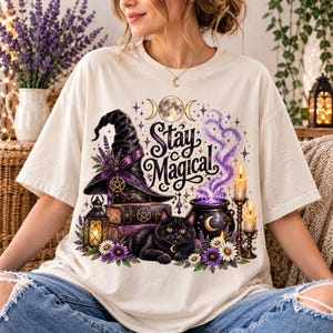 Könnte beinhalten: Ein cremefarbenes T-Shirt mit einem Grafikdruck, der eine schwarze Katze, einen Hexenhut, einen Bücherstapel, einen Kessel mit violettem Rauch, Kerzen und Blumen zeigt. Der Text "Stay Magical" ist in einer dekorativen Schriftart aufgedruckt.