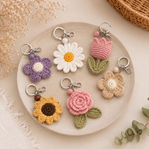 Puede incluir: Seis llaveros de flores de ganchillo que incluyen una margarita morada, una margarita blanca con centro amarillo, un tulipán rosa con hojas verdes, una flor beige, un girasol amarillo con centro marrón y una rosa rosa con hojas verdes.