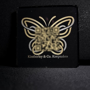 May include: A square black plaque featuring a gold and silver butterfly graphic with a blurred QR code centred on the wings. The text Kimberley & Co. Keepsakes is printed in gold lettering at the bottom of the plaque against the black background.