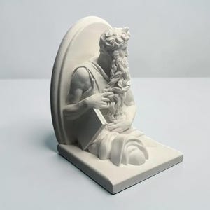 Könnte beinhalten: Ein weißer, matter Buchstützen-Skulptur einer bärtigen Person mit lockigem Haar, die in drapiertes Gewand gekleidet ist und ein rechteckiges Buch hält. Die Figur ist vor einer abgerundeten Rückwand auf einer flachen, rechteckigen Basis platziert.