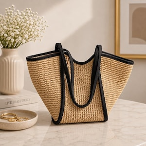 May include: A woven straw tote bag featuring a natural tan textured body and contrasting black fabric trim along the top edges and the two integrated shoulder straps. The bag has a wide, open-top design with a structured, curved silhouette.