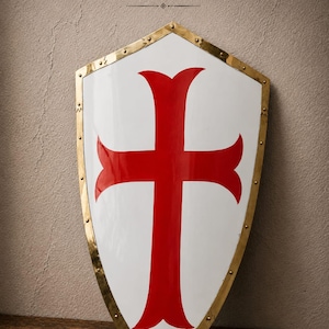 May include: A white shield featuring a large red cross in the centre. The shield has a metallic gold-coloured border secured with rivets along the edges. The text DORCAS CRAFT is printed in black above the shield on the wall surface.