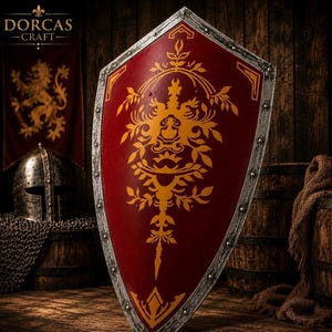 May include: A pointed heater shield featuring a deep red surface with a prominent gold-colored floral and scrollwork design. The shield has a metallic silver-toned border with visible rivets along the edge, creating a structured and durable appearance.