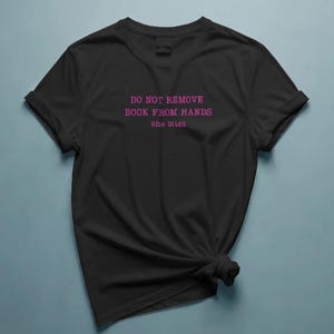 以下が含まれることがあります： クルーネックとロールアップされた袖が特徴の黒い半袖Tシャツ。胸の中央にはピンク色の文字で「DO NOT REMOVE BOOK FROM HANDS she bites」とプリントされています。Tシャツの右下は結び目になっています。