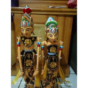 Puede incluir: Dos marionetas de varilla de madera wayang golek javanesas con rostros pintados a mano, tocados elaborados con detalles en rojo, azul y verde, y corpiños de terciopelo negro adornados con bordados florales dorados y cuentas sobre faldas de batik.