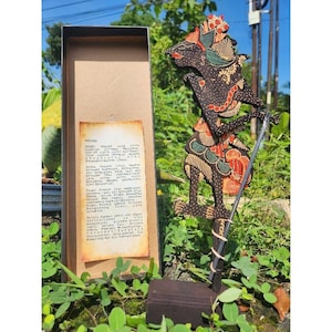 Könnte beinhalten: Eine javanische Wayang-Kulit-Schattenpuppe, bestehend aus einer dunkel geschnitzten Figur mit roten, grünen und goldenen Farbakzenten auf einem Holzsockel, neben einer rechteckigen Schachtel mit einer beschreibenden Karte in indonesischer Sprache.