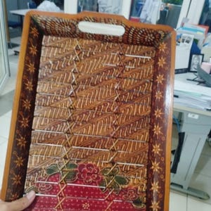 Pode incluir: Uma bandeja de servir retangular de madeira com um padrão batik castanho de linhas intrincadas e onduladas. A borda é decorada com um motivo de estrelas repetitivo. A seção inferior exibe um painel vermelho com flores e folhas verdes.