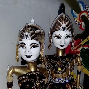 May include: Two Javanese wayang golek rod puppets featuring white faces with black painted features, gold-toned headpieces, and black velvet clothing adorned with gold beadwork and blue floral waistbands. The puppets have articulated arms.