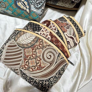 May include: A collection of three zippered pouches featuring intricate batik patterns in shades of brown, black, and cream. Each pouch has a curved top edge with a light-colored trim and a black zipper closure, showcasing detailed floral and swirl motifs.