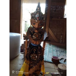 Pode incluir: Uma marioneta Wayang Golek com uma cabeça de madeira esculpida detalhada com rosto pintado, um toucado alto e pontiagudo, e um corpete de veludo preto adornado com missangas douradas. A marioneta usa um sarong estampado e fitas amarelas.