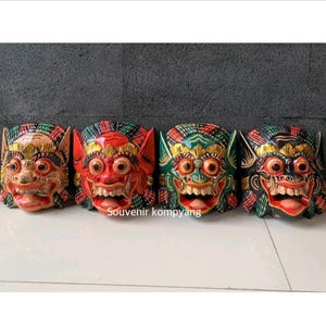Op de afbeelding: Vier handgesneden houten maskers op een rij, elk met een gestileerd gezicht met prominente ogen, ontblote tanden en gedetailleerde beschilderingen in beige, rood, groen en zwart. De tekst "Souvenir kompyang" is in het midden zichtbaar.