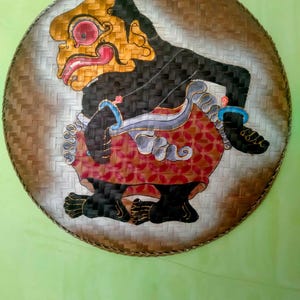 Op de afbeelding: Een ronde schaal van gevlochten bamboe met een handgeschilderde figuur met een zwarte huid, een rood patroon op het onderkleed en blauwe accenten. De figuur heeft een opvallende neus, een groot rood oog en een geopende mond.