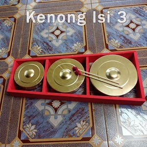 Puede incluir: Un conjunto de tres instrumentos kenong de metal dorado de diferentes tamaños, cada uno con una cúpula central elevada, alojados en un marco rectangular de madera roja. Dos mazos de madera con puntas acolchadas rojas descansan sobre ellos.