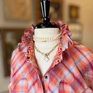 May include: A plaid button-down shirt in shades of orange, pink, and light blue, featuring a ruffled collar with a floral pattern on the underside. The shirt is styled with a layered necklace consisting of a pearl strand and a gold chain with a clover charm.
