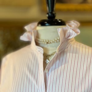 May include: A long-sleeved button-down shirt featuring a white and red vertical pinstripe pattern. The shirt has a high, ruffled collar and a front button placket. A single chest pocket is visible on the right side of the garment.