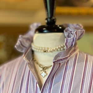 May include: A button-down shirt featuring a vertical striped pattern in shades of purple, white, and maroon, with a high ruffled collar. The shirt is styled with a multi-strand pearl necklace and a gold-tone chain necklace with a clover pendant.
