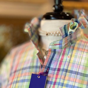 Peut inclure: Une chemise à manches longues boutonnée avec un motif à carreaux multicolores dans des tons de vert, bleu, rose et jaune. La chemise présente un col à volants et une étiquette rectangulaire bleue attachée par un cordon violet, portant le texte Alan Flusser.