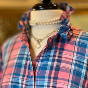 May include: A button-down shirt featuring a plaid pattern in shades of pink, blue, and white. The shirt has a ruffled collar, a front button placket with pink buttons, and a chest pocket. A layered necklace with pearls and a metal chain is visible.