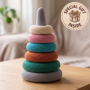 May include: A crochet stacking toy featuring six rings in shades of gray, cream, pink, teal, rust, and dark green, topped with a gray cone. A circular beige badge in the upper right corner displays a gift icon and the text SPECIAL GIFT INSIDE.
