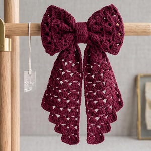 May include: A deep burgundy crochet bow featuring an intricate openwork pattern with two long, cascading tails. The bow is constructed from yarn and is displayed hanging from a light-coloured wooden stand, showcasing its textured, handmade design.