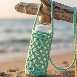 May include: A clear glass water bottle with a light brown cap, encased in a mint green crocheted sleeve featuring a diamond-shaped lattice pattern and a small decorative bow at the top, complete with a long matching crocheted carrying strap.