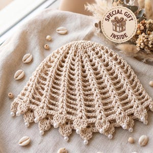 Puede incluir: Un sombrero de ganchillo beige con un patrón de abanico calado y un borde festoneado adornado con pequeñas cuentas blancas. El sombrero se muestra junto a conchas y cuentas de madera, con una etiqueta circular que dice SPECIAL GIFT INSIDE.